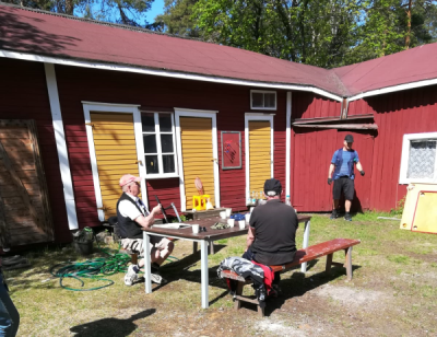 Vanhan maalaistalon aurinkoisella pihalla kaksi ihmistä kahvittelee pöydän ääressä, taustalla mies hanskat kädessä.