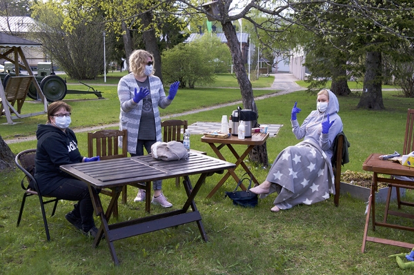 Kolme naista maskit naamalla ja kumihanskat kädessä vehreällä pihalla, jossa pihakalusteita aseteltu erilleen toisistaan.