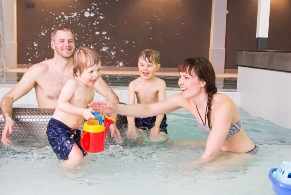Äiti, isä ja kaksi nuorto poikaa loiskuttavat iloisesti vettä sisäuima-altaassa.